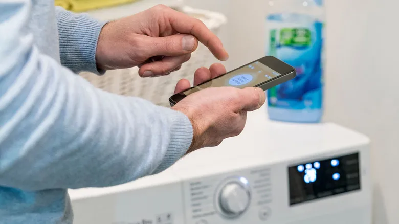 A man holding his phone in front of a washing machine.