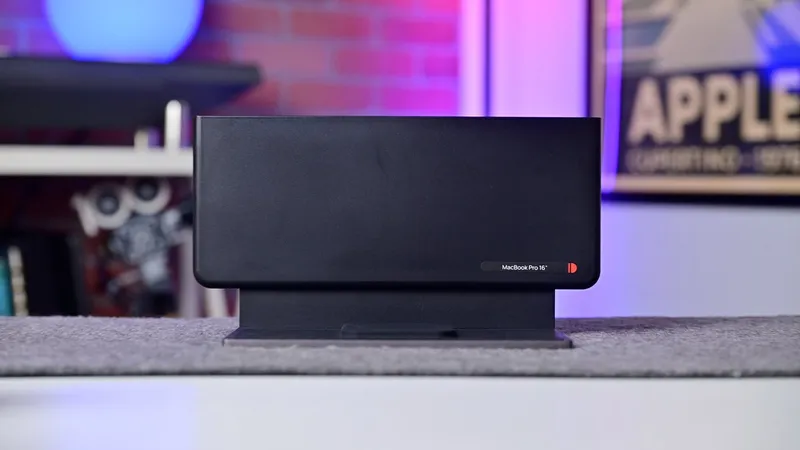Black rectangular electronic device on a stand, centered on a gray tabletop, with blurred shelves and colorful purple and pink lighting in the background