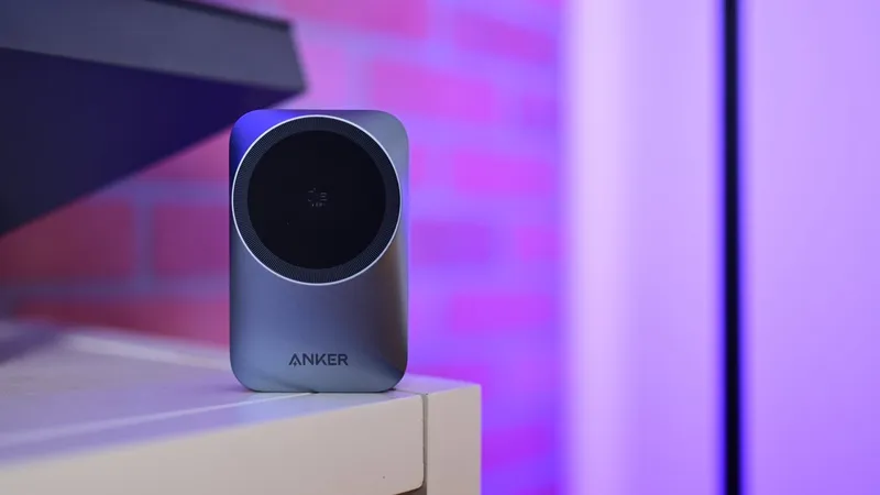Small silver Anker gadget with a circular black front sits on a light-colored table, with a softly lit purple and pink brick wall background
