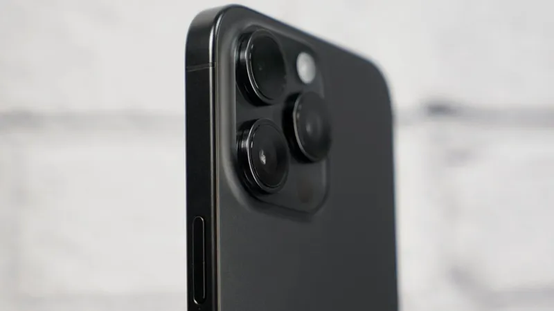 Close-up of an iPhone 15 Pro Max showing three large rear camera lenses and flash on a raised square module against a soft, out-of-focus light background