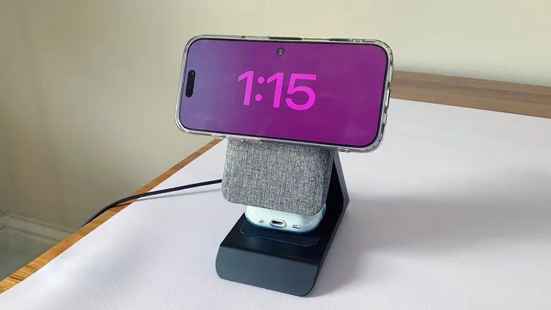 Smartphone in landscape mode displaying 1:15 with bright pink numbers, mounted on a gray fabric-covered charging stand holding wireless earbuds, on a white desk against a plain wall