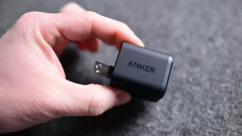 Hand holding a small black Anker wall charger with metal prongs extended, resting against a soft gray fabric background