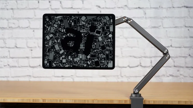 iPad Pro on adjustable metal arm above wooden desk, showing AI letters formed by many tiny gadget icons, against a white brick wall background.