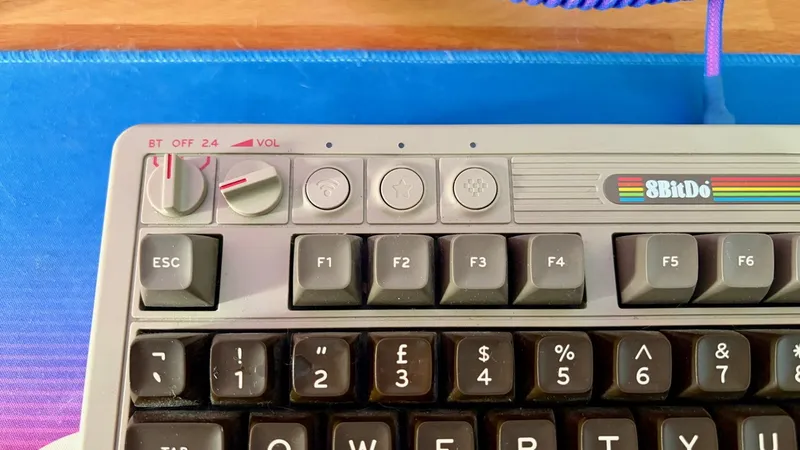 Retro-style 8BitDo keyboard close-up showing function keys, escape key, wireless and star buttons, round volume and mode knobs, on a blue desk mat with coiled cable above.