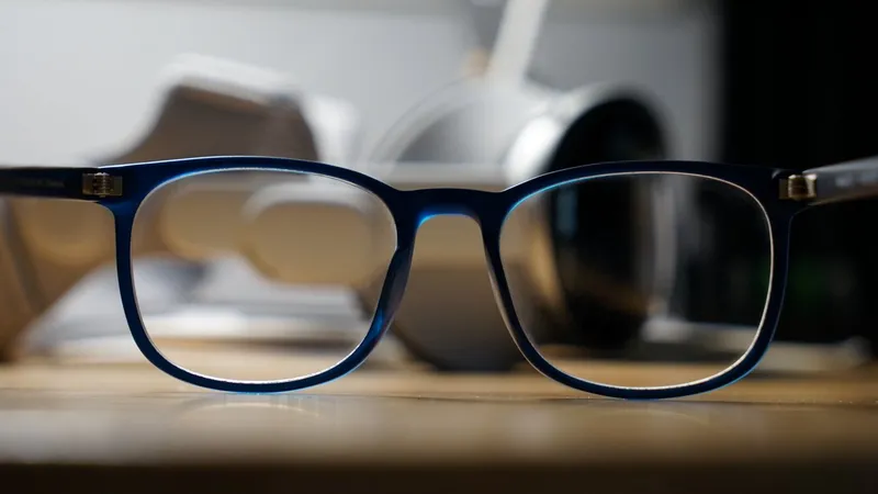 Blue rectangular eyeglasses resting on a wooden surface, sharply focused, with blurred white headphones and other desk objects in the background