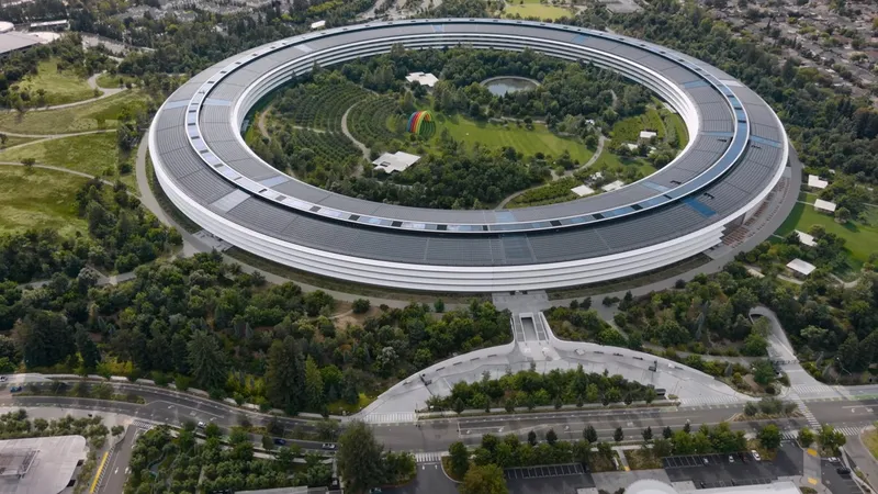 Aerial view of a Apple Park's Ring campus surrounded by dense green trees, inner courtyard park, pathways, and parking lots, set within a suburban landscape with roads and houses nearby