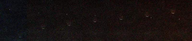 A grainy black image showing a horizontal sequence of seven faint crescent shapes evenly spaced from left to right. A tiny bright white dot moves diagonally from the upper left to the lower right, appearing to pass behind the crescent shapes.